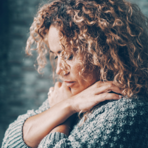 A person with long curly hair looks down and sad as she hugs herself.