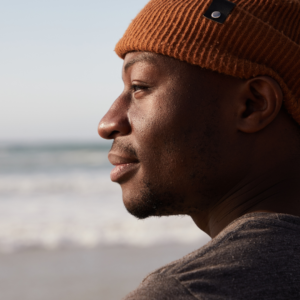 Black man with orange hat stares in the distance from profile with ocean in background. 